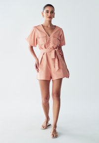 Persika värviga romper, millel on sügav V-kaelus, lühikesed puhvärved, kaks rinna taskut ja seotud vöökoharihm. Sile kangas ja vabalt langev lõige.