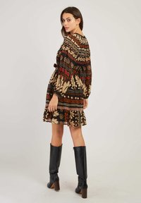 Femme vêtue d'une robe longue à manches longues à motifs, mélangeant des designs géométriques et floraux, associée à des bottes noires à talons montantes jusqu'aux genoux, se tenant de côté.