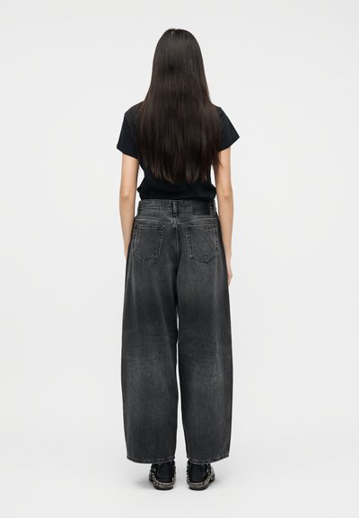 Γυναίκα με μακριά σκούρα μαλλιά στέκεται γυρισμένη πλάτη, φορώντας μαύρο μπλουζάκι, φαρδιά ξεβαμμένα μαύρα τζιν και μαύρα αθλητικά παπούτσια με μεταλλικές λεπτομέρειες.