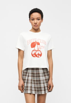 Femme portant un T-shirt blanc avec un motif de cerises rouges et le texte "Fruit Club", associé à une jupe plissée à carreaux marron et blanc.