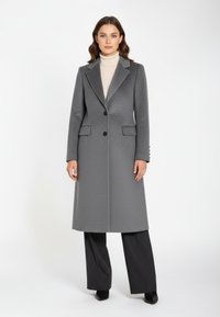 Grauer Wollmischmantel mit strukturiertem Design, aufgeschlagenen Revers, zwei Fronttaschen und schwarzen Knöpfen. Kombiniert mit dunklen Hosen und Stiefeletten.