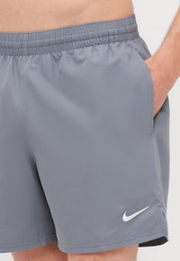 Shorts de sport gris avec une taille élastique, des poches latérales et un logo Nike blanc sur le bas à gauche. Texture lisse.