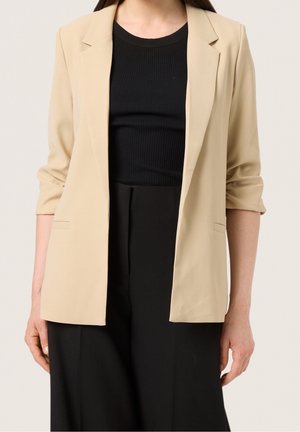 Kobieta ubrana w beżową marynarkę na czarnym, prążkowanym topie i czarnych spodniach, z marynarką z podwiniętymi rękawami do łokci.