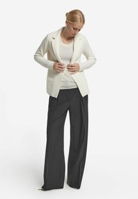 Elfenbeinfarbener Strickblazer mit Reverskragen, Fronttaschen und strukturiertem Muster, kombiniert mit dunklen, weiten Hosen und einem taillierten Oberteil.