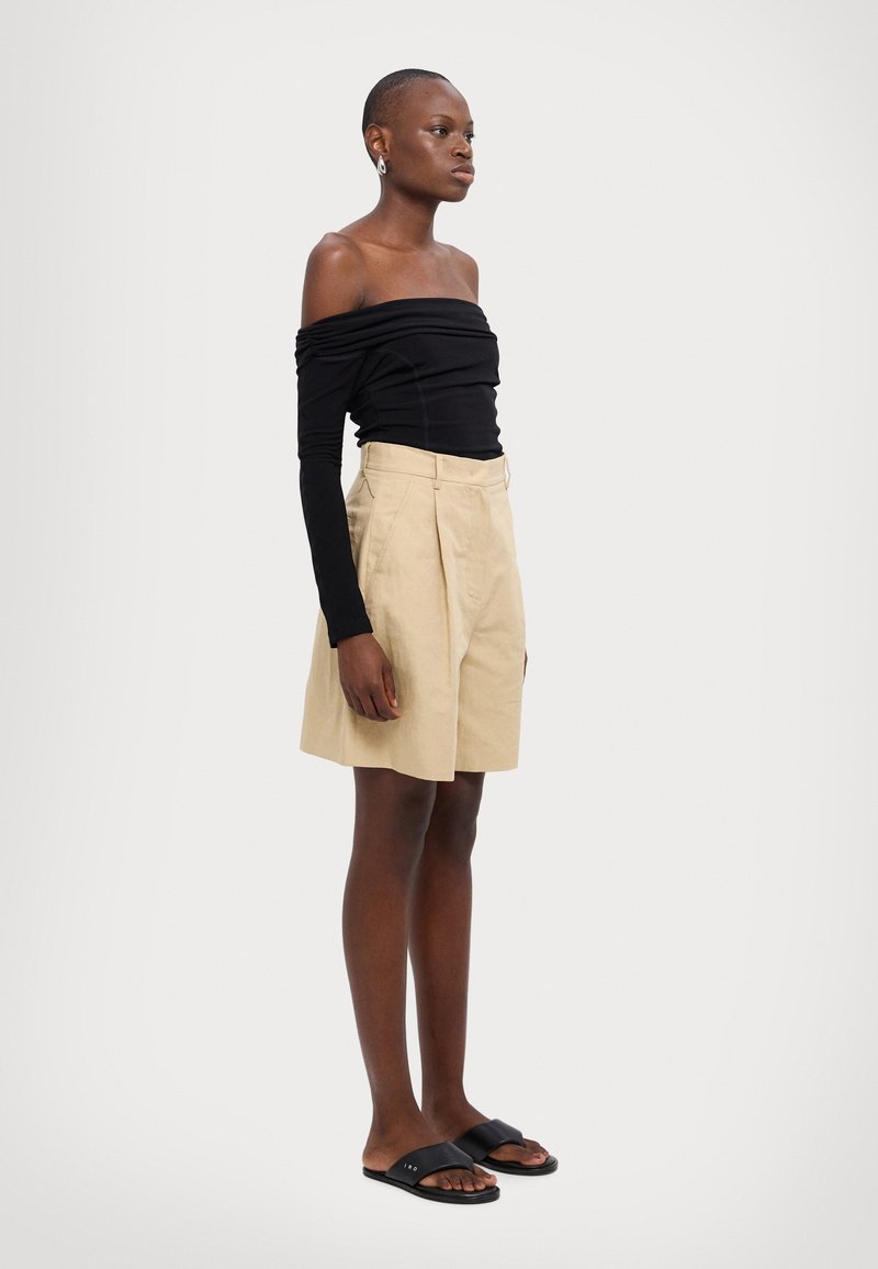 Schwarzes Off-Shoulder-Top mit langen Ärmeln, beigen gefältelten Shorts und schwarzen Rutschsandalen. Glattes Material und neutrale Farbpalette.