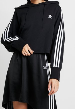 Personne portant un sweat à capuche noir Adidas et une jupe noire assortie avec des détails blancs à trois bandes et des logos Adidas sur les manches et la jupe.