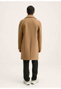 Kameelwollen jas met een getailleerde silhouet, geronde kraag en enkele achtersplit. Zwarte broek en lichte sneakers maken de look compleet.