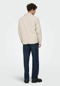 Veste-chemise beige clair avec col pointu, poignets à boutons et coupe droite, associée à un jean bleu foncé à jambes droites.