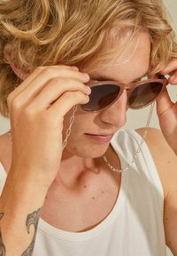 Light brown sunglasses with dark lenses, a metal chain connecting to the frame, worn by a person with tousled blonde hair.