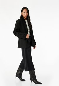 Blazer negro de gran tamaño sobre una camiseta de rayas blancas y negras, combinado con pantalones anchos negros y botas de tacón alto negras hasta la rodilla. Textura suave.