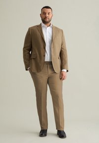 Un costume en lin marron clair avec une veste à deux boutons, porté sur une chemise blanche. Coupe ajustée, pantalon à jambe droite, associé à des chaussures noires.