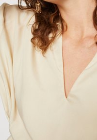 Haut en tissu doux de couleur crème avec un décolleté en V, une texture lisse et une coupe décontractée avec des accents de manches froncées.