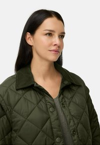 Vrouw met lang, donker haar, gekleed in een donkergroene gewatteerde jas over een grijze gebreide top, die iets opzij kijkt tegen een effen achtergrond.
