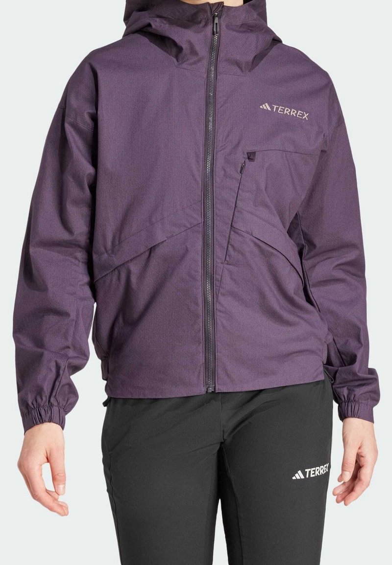 Personne portant une veste zippée Terrex violet avec capuche et un pantalon Terrex noir, debout les mains détendues le long du corps.