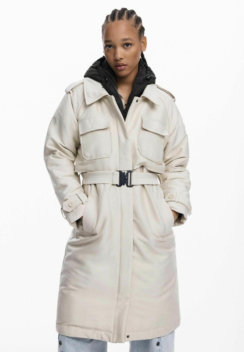 Trench coat de culoare bej deschis, cu talie subțire, două buzunare pe piept și un căptușel cu glugă neagră. Prezentă o textură netedă și manșete cu nasturi.