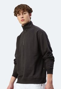 Dunkelgrauer Pullover mit hohem Kragen, halbem Reißverschluss an der Vorderseite, langen Ärmeln und seitlichen Belüftungen. Hergestellt aus weichem Stoff mit glatter Textur.