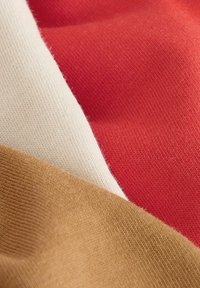 Primo piano di strati di tessuto morbido nei colori marrone cammello, panna e rosso con trama a maglia visibile.