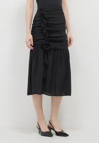 Frau in einem schwarzen Midirock mit Rüschendetails und schwarzen Slingback-Pumps, die an einer hellen Wand steht.