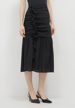 Femme portant une jupe midi noire avec des détails froncés et des escarpins à talon noir, se tenant contre un mur clair.