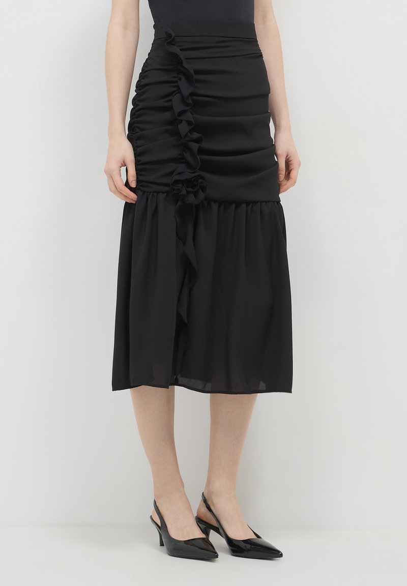 Frau in einem schwarzen Midirock mit Rüschendetails und schwarzen Slingback-Pumps, die an einer hellen Wand steht.