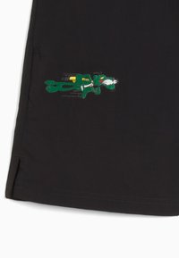 Zwarte shorts met een groene geborduurde cartoonfiguur in beweging, met rode ogen en een gele accent op zijn rug.