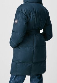 Mörkblå puffjacka med bälte i midjan, kviltat mönster och en rund logomärke på vänster ärm. Mjuk, slät struktur.