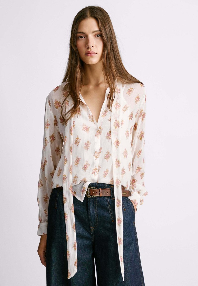 Blusa floreale in tessuto leggero e semi-trasparente, con motivi floreali rosa, chiusura frontale con bottoni e maniche lunghe, abbinata a pantaloni di jeans a vita alta.