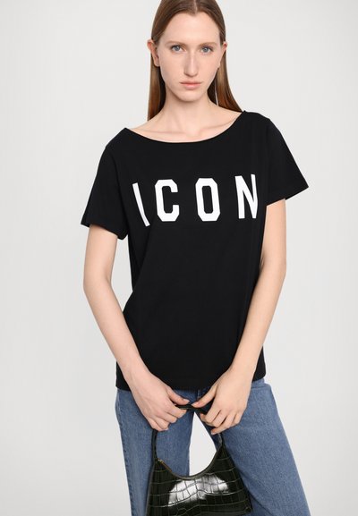 T-shirt en coton noir avec un graphique blanc "ICON", coupe ample, col large, associé à un jean bleu et un sac en cuir verni vert.