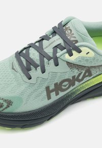 La chaussure de course HOKA Challenger ATR présente un upper en mesh vert clair, des lacets gris foncé, une semelle texturée et des accents vert fluo.
