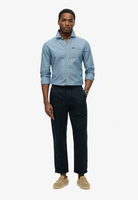 Camicia chambray azzurro chiaro, pantaloni scuri con coulisse e scarpe beige slip-on. Le caratteristiche includono una tasca sul petto e maniche arrotolate.
