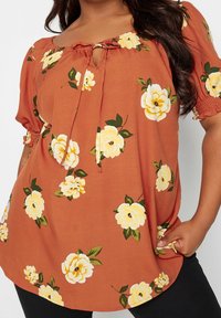 Bluzka w kolorze rdzy z kwiatowym wzorem w żółtym i zielonym kolorze. Posiada dekolt w kształcie litery U oraz elastyczne rękawy z wiązanym detalem z przodu.