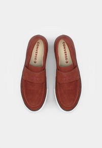 Sapatos slip-on em camurça vermelha com biqueira arredondada, sola de borracha branca e um destaque em forma de estrela perto da alça. Logótipo "CONVERSE" impresso no interior.