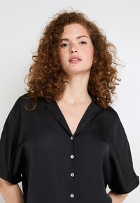 Chemise noire à manches courtes avec une coupe décontractée, fabriquée en tissu lisse, dotée d'un col et d'une fermeture à boutons à l'avant avec des boutons de teinte claire.