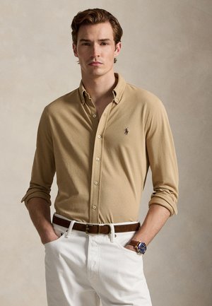 Férfi bézs hosszú ujjú ingben, amihez logó tartozik, fehér nadrágban, barna övvel és kék karórával, zsebében tartja a kezét.