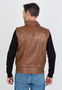 Braune, ärmellose Lederweste mit einem spitzen Kragen, Rückennähten und einem verstellbaren Riemen an der Taille. Getragen über einem schwarzen Shirt.