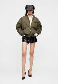 Γυναίκα που φοράει μπουφάν bomber σε πράσινο ελιάς με επένδυση, μαύρα σορτς από δερματίνη, ψηλά τακούνια και γκρι καπέλο με μαύρα γούνινα καλύμματα αυτιών.