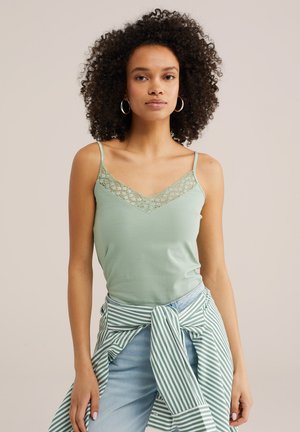 Žena kučeravých vlasov, oblečená v svetlozelenej blúzke s čipkovou bordúrou, kruhových náušniciach, svetlomodrých džínsach a zelenej pruhovanej košeli zviazanej okolo pása.