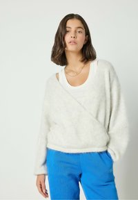 Weißer, flauschiger Pullover mit tiefem V-Ausschnitt und langen Ärmeln, über einem weißen Tanktop getragen. Kombiniert mit blauen, strukturierten Hosen.