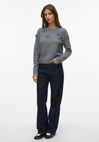 Frau in einem grauen Pullover mit floralen Applikationen, dunkelblauen, weit geschnittenen Jeans und schwarz-weißen Slippern, stehend vor einem weißen Hintergrund.