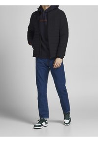 Schwarze gepolsterte Jacke mit Kapuze, getragen über einem dunklen Hoodie mit orangefarbigem Text. Kombiniert mit blauen Jeans und schwarz-weißen Sneakers. Modernes Design.
