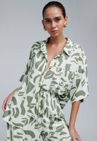 Camicia verde chiaro con bottoni, decorata con motivi astratti, maniche corte e tasca, abbinata a pantaloni con cordoncino coordinati. Tessuto texturizzato.