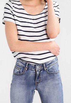 Personne portant une chemise blanche à manches courtes avec des rayures horizontales noires et un jean bleu en denim, les bras croisés, sur un fond uni.