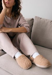 Beige, flauschige Hausschuhe aus weich material, mit runder Zehenform und strukturierten Seiten, getragen mit weißen Socken, vor einem hellen Sofa.