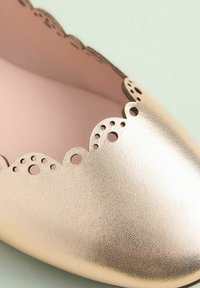 Goldene Lederflats mit gewelltem Rand und runden Ausschnitten sowie einer glatten, strukturierten Oberfläche. Das Innere ist hellpink.