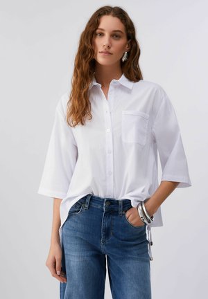 Frau mit langem, welligem Haar trägt ein lockeres, weißes Hemd mit Knöpfen und Tasche sowie eine blaue Jeans. Sie posiert mit einer Hand in der Tasche und silbernen Armbändern am Handgelenk.