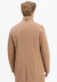 Tan lange jas met een gestructureerde kraag en een gladde textuur. Heeft naaddetails langs de achterkant en een logo bij de nek.