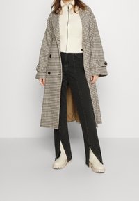 Long plaid coat with double-breasted buttons, layered over a cream sweater and black jeans with side slits. Beige ankle boots complete the look.