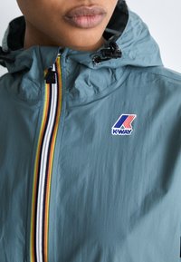 Chaqueta impermeable de color azul claro con capucha, que presenta un cierre vertical de varios colores y un logo de K-WAY en el lado izquierdo. Textura suave.