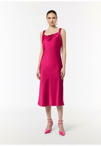 Różowa satynowa sukienka z dekoltem typu cowl, dopasowanym górą i sylwetką w kształcie litery A. Modelka łączy ją z pasującymi wysokimi sandałami na szpilce.
