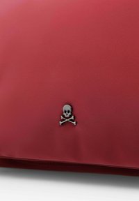 Funda roja texturizada con un pequeño emblema de calavera y huesos cruzados metálico. Exterior suave con un cierre de cremallera a lo largo del borde inferior.